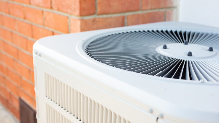 Close-up of a residential outdoor air conditioning condenser unit with visible fan blades, installed next to a red brick wall on a sunny day